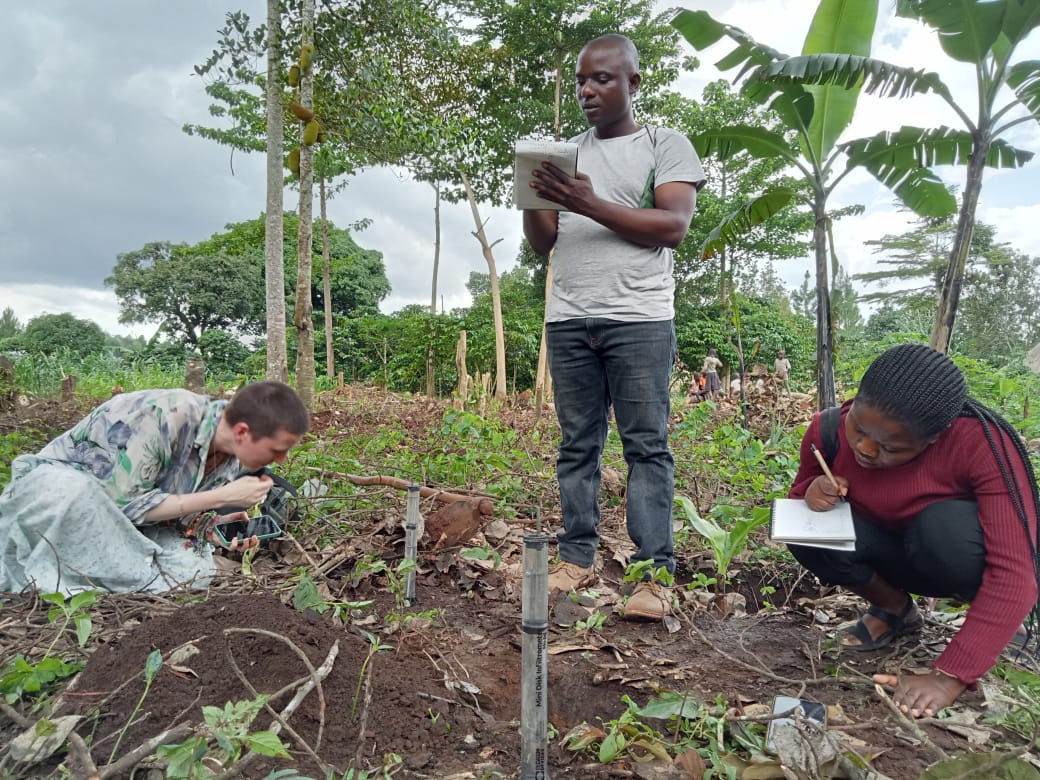 Cephas Chisanga, Rising Scholars course participant. The photo shows 1 person standing making notes on paper, and 2 people sitting on the ground looking at vegetation. The people are outside, with trees in the background, and small vegetation and earth in the foreground. 
