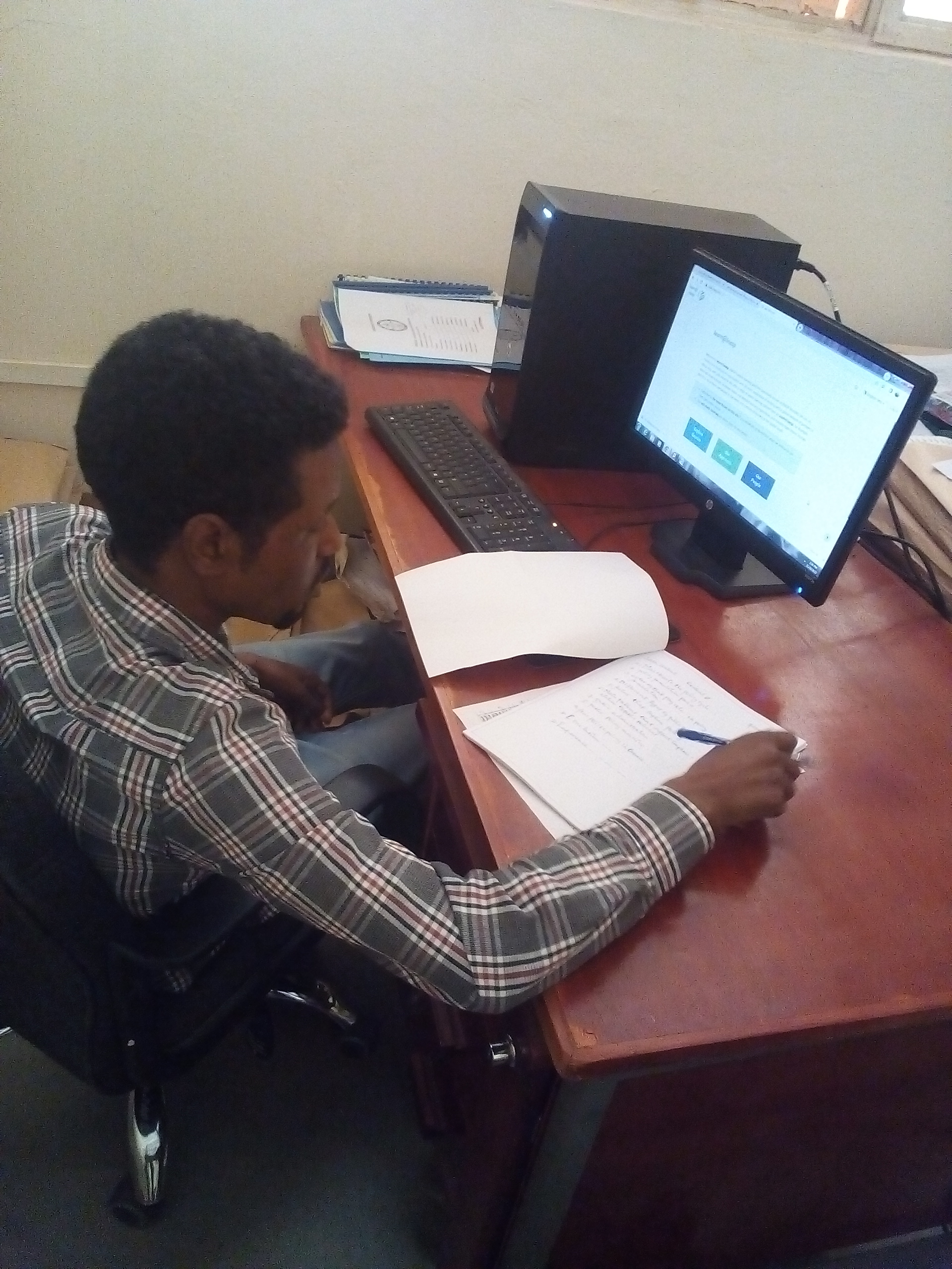 Mussie Gebreagziabher, a Rising Scholars course participant, sits at a desk, with papers, a computer, monitor, and keyboard on the desk. Mussie holds a pen and is looking at the papers and monitor screen.