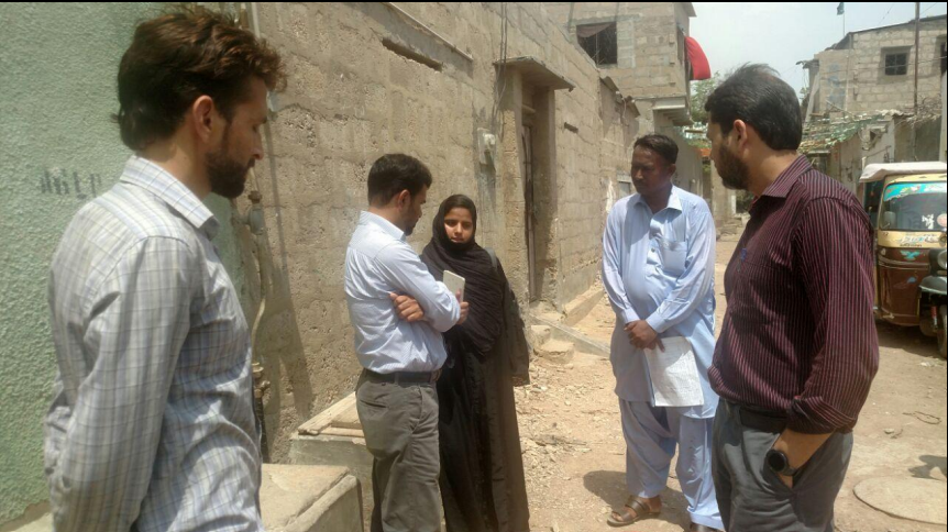 Five people standing on a street. Two of the people appear to be talking to each other, whilst the other three watch on. In the background are buildings on the left and right, and vehicles in the background on the right.