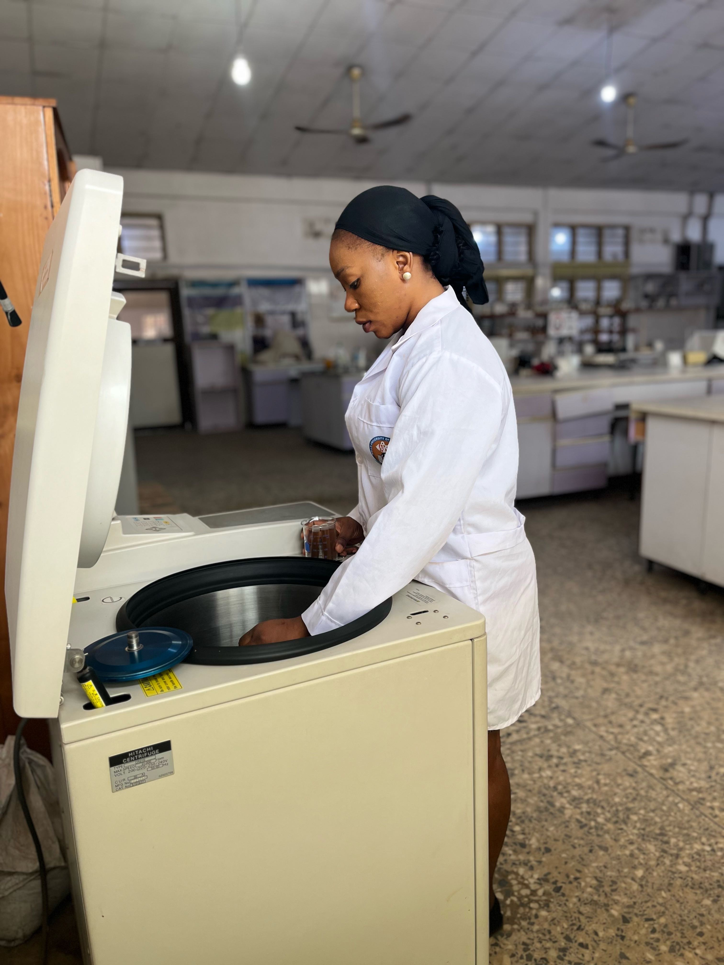 A photo showing a large machine on the left with an open lid. A person wearing a white lab coat stands at the machine, holding something obscured from view in the top of the machine. In the person's visible hand, they hold a glass beaker. The background shows a laboratory setting.