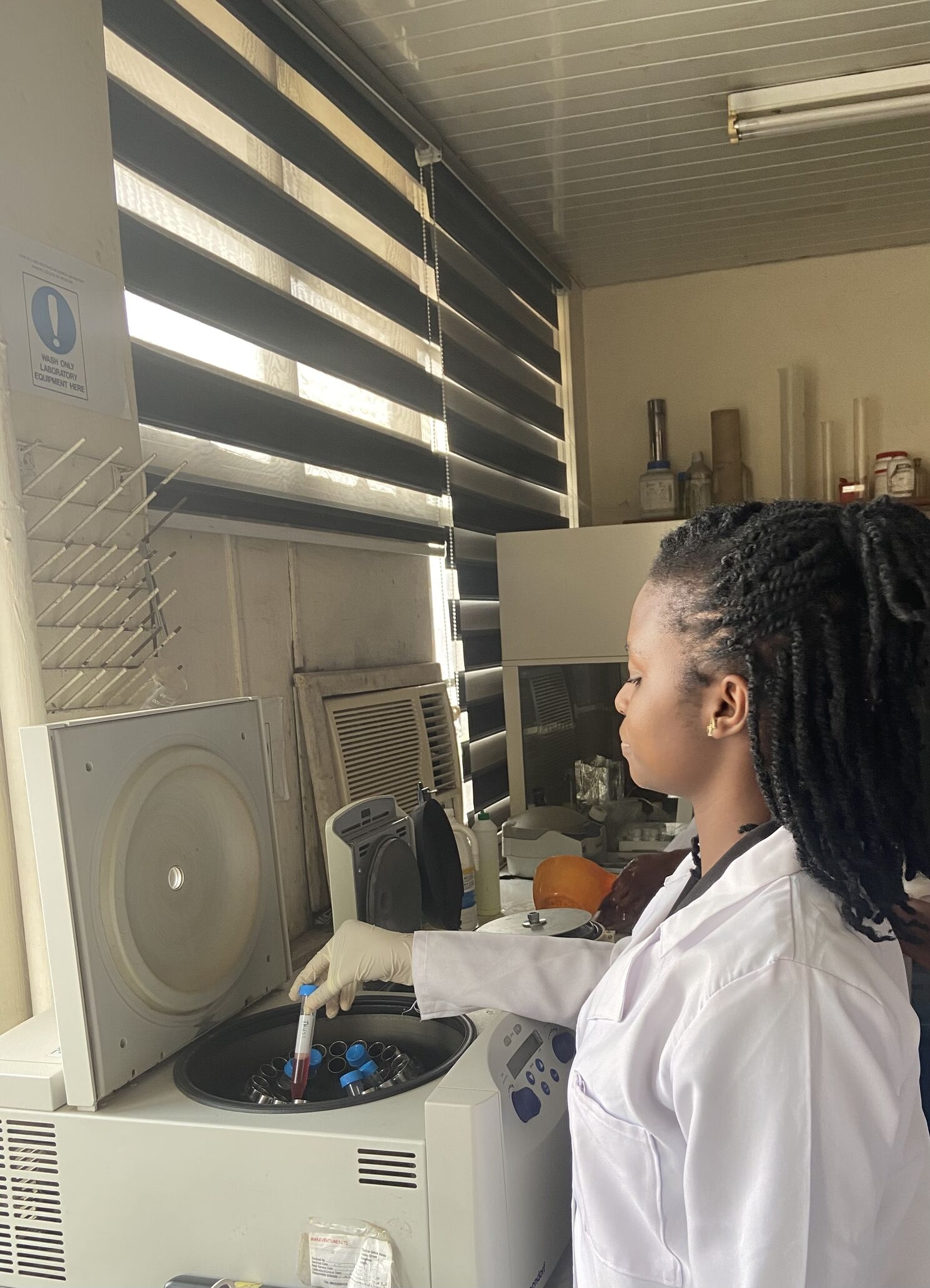 The photo shows a laboratory setting. A small machine is in the left of the image with an open lid. A person wearing a white lab coat and gloves holds a testtube with a blue lid in the machine. 