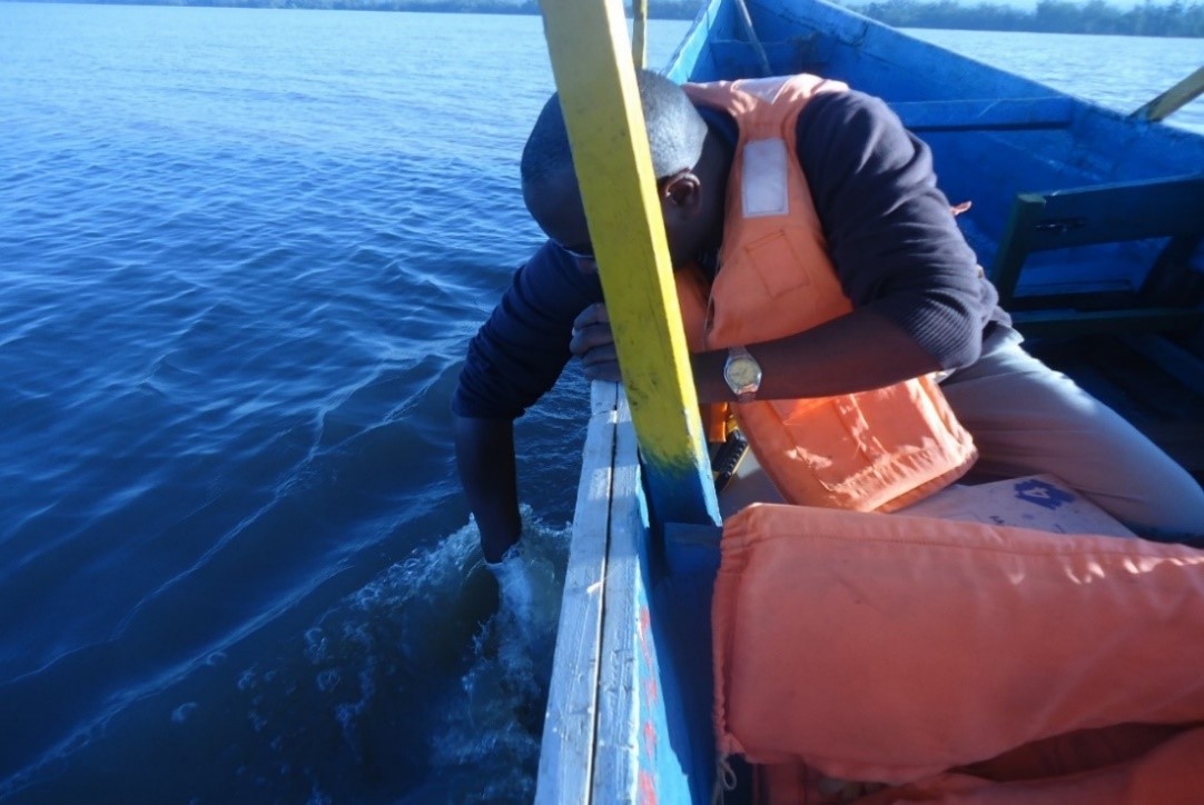 The photo shows a large body of water, and on the right side of the image, a person wearing an orange lifevest sitting in a wooden boat and leaning over the side holding something in the water.